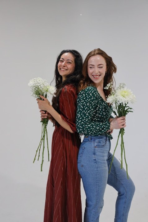 Two women standing back to back holding bouquets of white flowers and laughing.